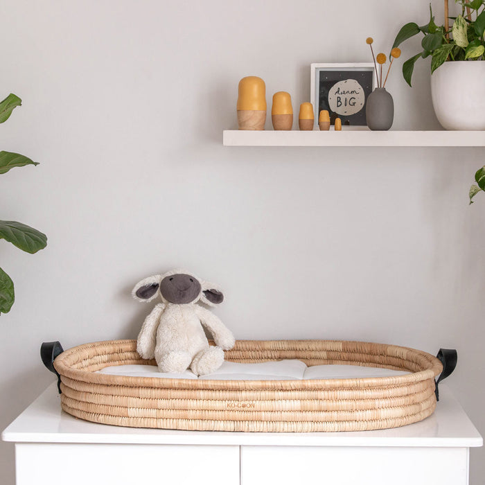 A wicker baby changing basket with leather handles, placed on a white surface, with a plush toy sheep inside. The basket is situated against a backdrop of a plant, shelves with decorative items, and a window.