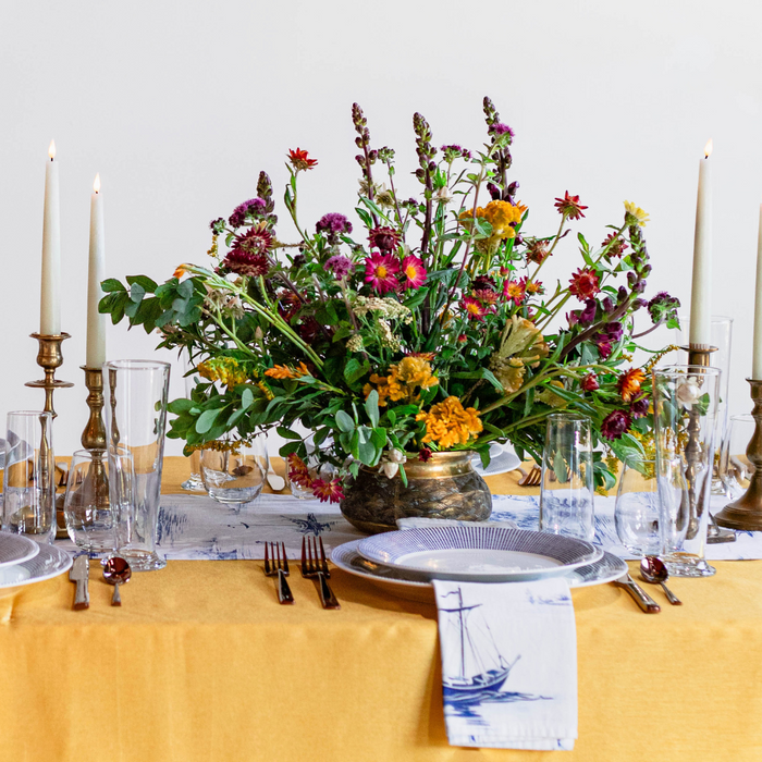 A table setting with blue and white dishes, rose gold flatware, casual stemless glassware and pint glasses, and a floral centerpiece, arranged on a yellow tablecloth.