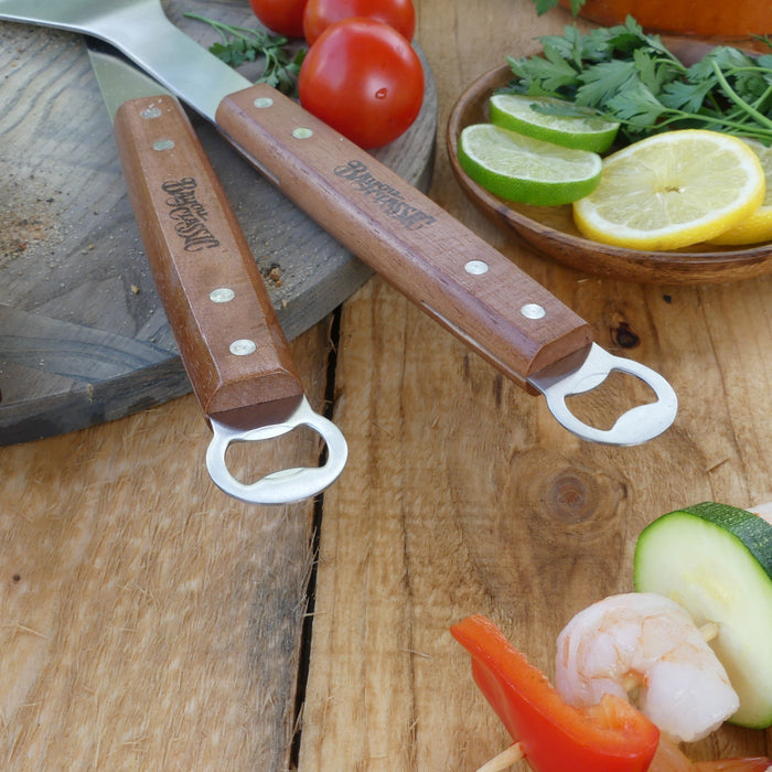 The bottle opener end of the spatula and tongs on a wood surface with vegetables.