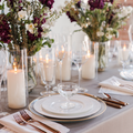A set table with event rentals including white rimmed dinnerware, rose gold flatware, and glassware including champagne flutes and red wine glasses, adorned with ivory satin and faux dupioni linen napkins, and floral arrangements with battery candles in the background.