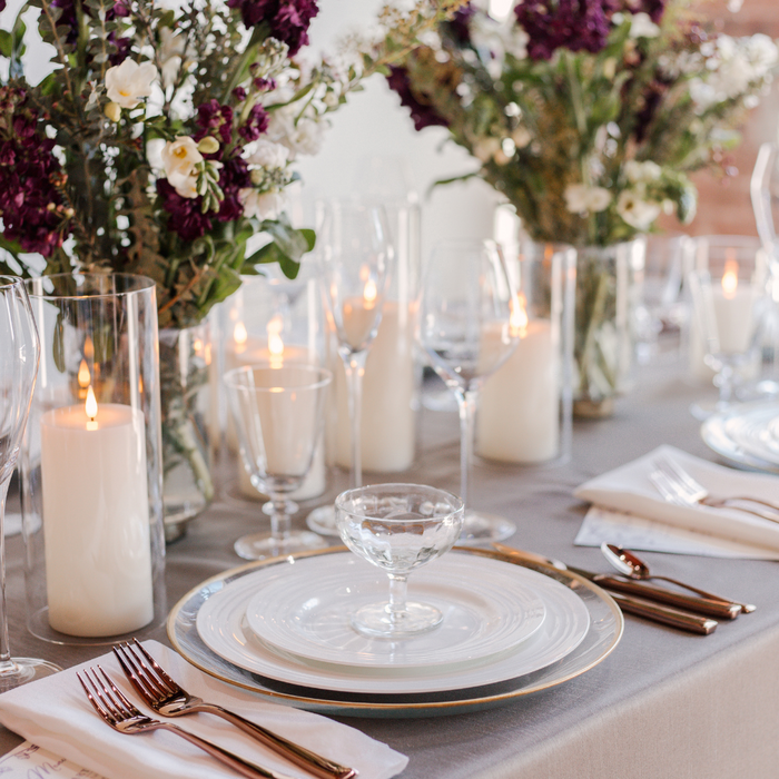 A set table with event rentals including white rimmed dinnerware, rose gold flatware, and glassware including champagne flutes and red wine glasses, adorned with ivory satin and faux dupioni linen napkins, and floral arrangements with battery candles in the background.