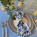 A set table with blue floral batik linen napkin, blue dotted dinnerware, vintage silverplate flatware, and etched glassware on a sky blue tablecloth.