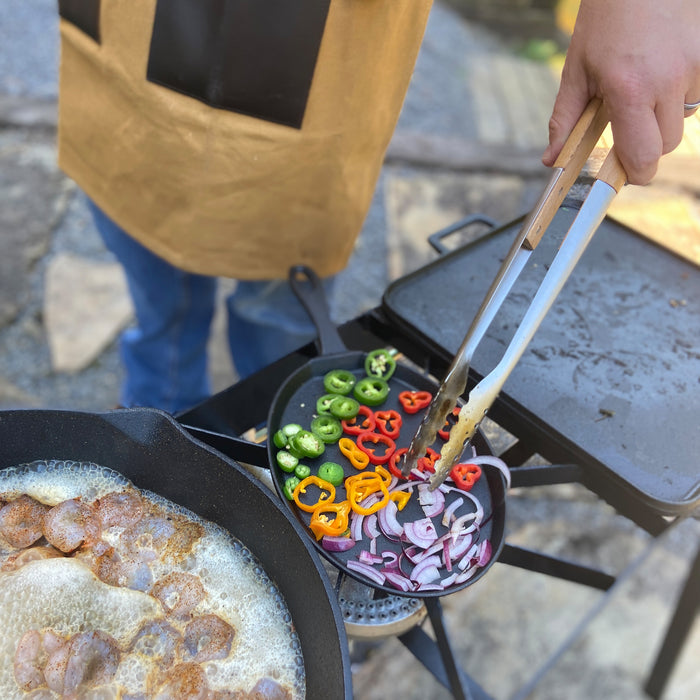 man holding grill tongs cooking vegetables and shrimp outside on an outdoor cooker